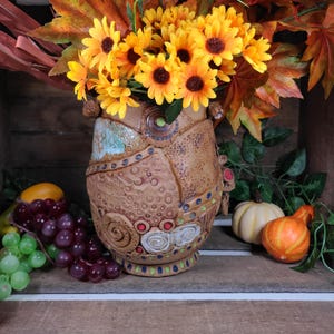 May include: A decorative ceramic vase with a textured, brown surface, adorned with colorful accents and filled with bright yellow sunflowers. The vase is surrounded by fall foliage, grapes, and pumpkins, creating a seasonal display.