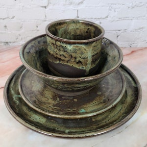 Puede incluir: Un juego de vajilla de cerámica hecho a mano con un esmalte marrón oscuro y verde. El juego incluye una taza, un cuenco y dos platos. El esmalte tiene un aspecto moteado, y los bordes de los platos y el cuenco están ligeramente elevados.