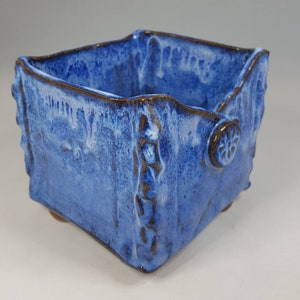 Pu&ograve; includere: Un vaso quadrato in ceramica fatto a mano con una glassa blu strutturata. Il vaso ha un bordo marrone e dettagli decorativi, tra cui un piccolo emblema circolare su un lato. Il vaso poggia su piccoli piedini.
