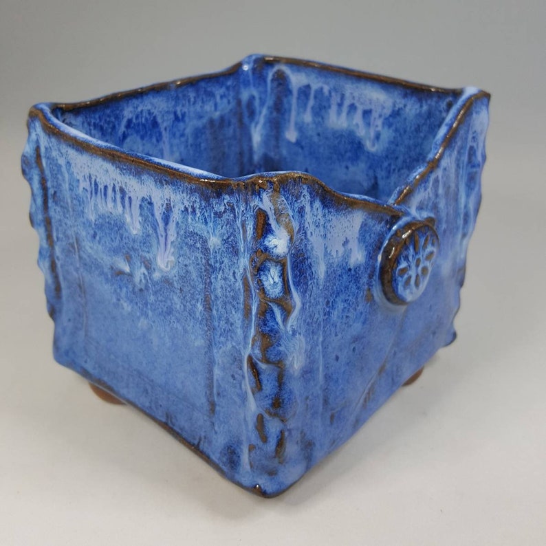 Pu&ograve; includere: Un vaso quadrato in ceramica fatto a mano con una glassa blu strutturata. Il vaso ha un bordo marrone e dettagli decorativi, tra cui un piccolo emblema circolare su un lato. Il vaso poggia su piccoli piedini.