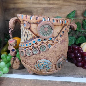 May include: A handcrafted ceramic pot with a textured, earthy brown surface. The pot features intricate designs with turquoise and green accents, including spiral and circular patterns. Decorative elements hang from the handles. The pot is approximately 7 inches tall.
