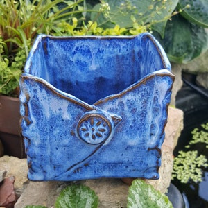 Pu&ograve; includere: Un vaso quadrato in ceramica blu con un design unico a forma di busta. Il vaso presenta una superficie strutturata con bordi marroni e un dettaglio a bottone decorativo. Ideale per piccole piante o succulente.