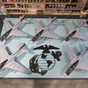 May include: Six gray and white model airplanes with red and black markings are arranged in a circle on a blue and white grid surface. A black metal emblem of the United States Marine Corps is in the center of the circle.