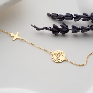 May include: A delicate gold necklace featuring a small airplane charm and a globe pendant. The necklace is displayed on a white surface with sprigs of lavender in the background. The chain is fine and the charms are detailed.