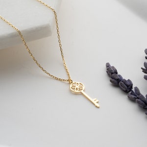 May include: A gold key necklace with a delicate chain and a detailed key pendant. The key has a floral design and a small loop at the top.