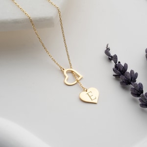 May include: A gold-colored necklace featuring two heart pendants. One heart is a cutout, and the other is a solid heart with the letter "E" engraved on it. The necklace hangs on a delicate chain. Lavender sprigs are in the background.