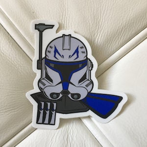 May include: A white and blue Star Wars clone trooper helmet sticker with a blue cape and a black and white blaster. The helmet has a blue stripe across the front and a white stripe across the back. The helmet has a white and blue design on the front.