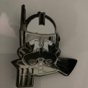 Può includere: Uno spillo in smalto nero e bianco con un teschio stilizzato che indossa un elmetto e una maschera antigas. Lo spillo è una rappresentazione di un clone trooper di Star Wars.