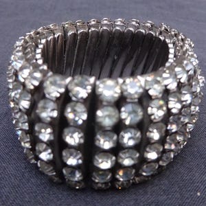 Puede incluir: Un brazalete plateado adornado con numerosos brillantes transparentes. El brazalete está sobre una tela gris oscuro, con una pared de ladrillos al fondo. Un accesorio audaz para ocasiones especiales.