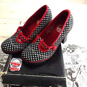 Puede incluir: Zapatos de lunares blancos y negros con ribetes rojos y una correa con botón rojo. El forro interior es rojo y la plantilla presenta el logotipo de Underground England con un gráfico de la Union Jack. Los zapatos descansan sobre una caja negra.