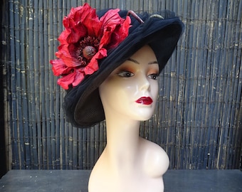 May include: A black wide-brimmed hat featuring a large, textured red poppy flower accent on the side. The hat is constructed from a dark, semi-sheer fabric with a structured brim and is displayed on a smooth, pale mannequin head with red lips.