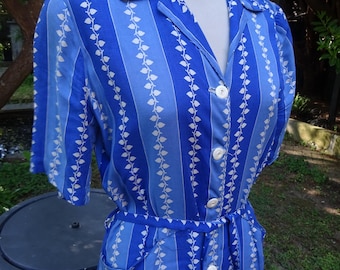 May include: A vintage blue dress with vertical stripes and a white leaf pattern. The dress features a collared neckline, short sleeves, and a button-down front. It has a matching belt and pockets. The dress is displayed on a mannequin.