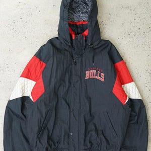 Peut inclure: Veste à capuche noire, rouge et blanche avec le logo des Chicago Bulls sur le devant. La veste a une fermeture éclair et une capuche à cordon.