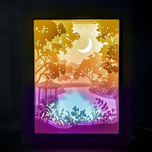 Peut inclure: Une œuvre d'art en papier en couches représentant un paysage serein avec un belvédère, un lac, des arbres et un croissant de lune. L'œuvre présente un dégradé de couleurs, passant du violet au bleu, puis à l'orange et au jaune.