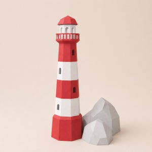 May include: A geometric paper lighthouse model featuring alternating horizontal red and white bands, a red roof, and a white lantern room with arched openings. Beside the lighthouse are two light gray, faceted, rock-like paper structures.