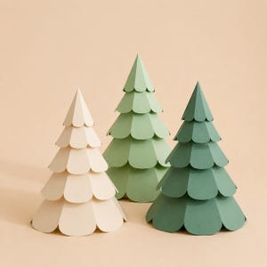 Könnte beinhalten: Drei konische Papierbäume in einer Gruppe angeordnet. Die Bäume haben geschichtete, bogenförmige Kanten, die an Tannennadeln erinnern. Ein Baum ist cremefarben, einer ist hell salbeigrün und der dritte ist in einem dunklen Waldgrün.
