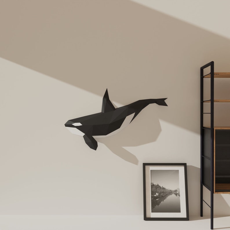May include: A black and white origami orca whale wall decoration. The orca is in a swimming position with its dorsal fin raised. The orca is mounted on a white wall.