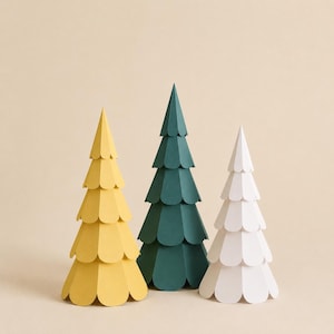 May include: Three conical paper trees arranged in a row. The left tree is yellow, the centre tree is dark green, and the right tree is white. Each tree features multiple horizontal tiers of rounded, petal-like paper cutouts stacked to form a layered structure.