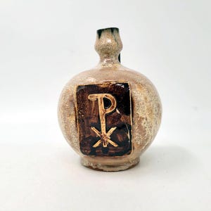 Op de afbeelding: Een ronde keramische vaas met een smalle hals en een textuurrijke, gespikkelde beige en bruine glazuurlaag. Een donkerbruine rechthoekige plaquette is in het midden aan de voorzijde geplaatst, met een lichtgekleurd Chi-Rho-symbool in reliëf.