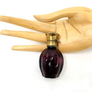 Puede incluir: Un frasco de perfume pequeño y redondo de vidrio morado oscuro con estrías verticales y una tapa metálica de color latón. El frasco presenta un cuerpo redondeado que se estrecha hacia el cuello, rematado con un tapón cilíndrico de metal liso.