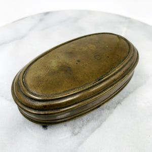 May include: An antique brass oval box with a hinged lid. The box has a tarnished, aged appearance, with a slightly textured surface. The lid and base are both oval, with a series of concentric rings adding detail to the design. The box is likely used for storing small items.