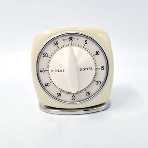 Puede incluir: Un temporizador de cocina Kienzle color crema con una esfera blanca y una base plateada. El temporizador tiene una perilla blanca y números negros que indican los minutos. La palabra "SIGNAL" está impresa en la esfera.
