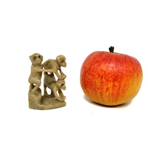 May include: A small, beige sculpture of three monkeys stacked on top of each other, next to a red and yellow apple. The sculpture is detailed, and the apple has a green stem.
