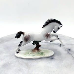 Op de afbeelding: Een witte keramische paardenfiguur met bruine vlekken en een donkere manen en staart. Het paard staat op zijn achterpoten, bevestigd aan een kleine boom en een groene basis. Decoratief item.