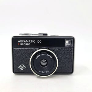 Puede incluir: Una cámara Agfamatic 100 negra con una lente plateada con la etiqueta "Agfa Colorstar Parator". La cámara tiene un punto rojo con la palabra "sensor" impresa en ella.