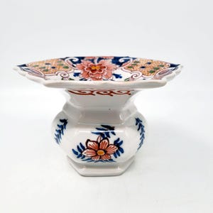 May include: A white ceramic vase with a wide, scalloped top and a flared base. The vase is decorated with floral patterns in shades of red, blue, orange, and green. The design is symmetrical and detailed, with a classic aesthetic.