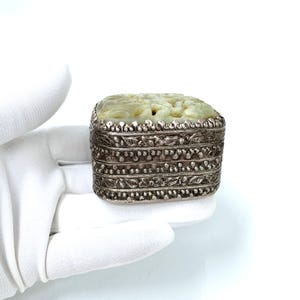 May include: A rectangular box featuring a pale green carved stone lid set into a silver-toned metal base. The metal sides are decorated with intricate embossed floral patterns and beaded borders, creating a textured, metallic finish.