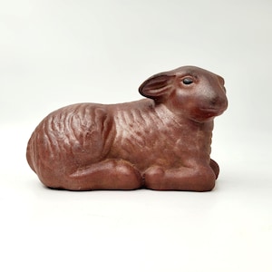 May include: A brown ceramic figurine of a lamb lying down. The lamb has a textured surface and black eyes.