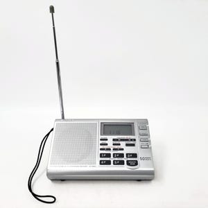 Puede incluir: Radio Sony plateada con antena extendida. La radio tiene una pantalla digital que muestra la hora. La parte frontal de la radio tiene una rejilla de altavoz y varios botones. Un cable negro está conectado al lateral.