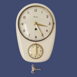 Op de afbeelding: Een crèmekleurige wandklok met een glanzende afwerking, voorzien van een ronde wijzerplaat met goudkleurige wijzers en cijfers, en een onderste ronde timer-wijzerplaat. De merknaam Mauthe staat op de wijzerplaat. Een kleine metalen opwindsleutel ligt eronder.