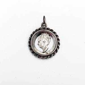 May include: A silver pendant featuring a profile of a person within a circular frame. The pendant has a twisted rope-like border and a small loop for a chain. The design is detailed and the metal has a slightly aged appearance.