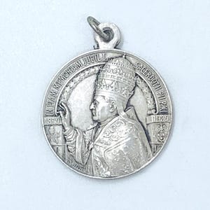 Op de afbeelding: Een zilverkleurige ronde hanger met een gedetailleerd reliëf van een religieuze figuur met een hoge hoed. De hanger heeft tekst rond de rand, inclusief de jaren 1879 en 1929. Bovenop zit een kleine lus.