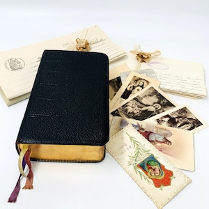 May include: A black leather-bound book with gold edges, alongside vintage postcards and envelopes. The book has colourful ribbon bookmarks. The postcards feature religious imagery. The envelopes are cream-coloured with faded text and stamps.