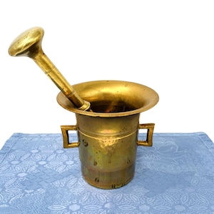 Op de afbeelding: Een vintage messing vijzel en stamper set. De vijzel heeft een brede, uitlopende rand en twee vierkante handvatten. De stamper is cilindrisch met een afgeronde kop. De set wordt gepresenteerd op een blauw patroon doek.