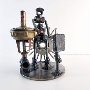Può includere: Una scultura in metallo fantasiosa di una figura realizzata con parti riutilizzate, che tiene una piccola valvola in ottone. La figura è posizionata su una base con un design a forma di ingranaggio.