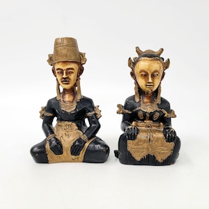 Op de afbeelding: Twee zittende beeldjes met crèmekleurige gezichten en zwarte lichamen met goudkleurige accenten. Het linkerfiguur draagt een hoog, cilindervormig gouden hoofddeksel, terwijl het rechterfiguur een versierde, meerpuntige gouden hoofdtooi heeft. Beide dragen goudkleurige kleding.