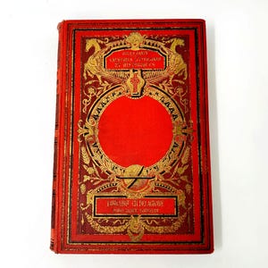 Puede incluir: Un libro antiguo con una cubierta roja y detalles dorados en relieve. La cubierta presenta diseños ornamentados, texto y un espacio circular central. El lomo del libro es visible, y la estética general sugiere un tema histórico o literario.