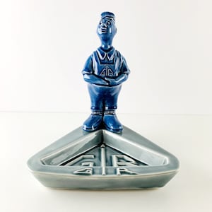 Puede incluir: Una figurita de cerámica azul de un hombre de pie sobre un cenicero triangular gris. El cenicero tiene las letras "G.R." en relieve en la parte inferior.