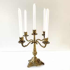 May include: A five-arm brass candelabra featuring ornate, embossed floral and leaf detailing on the base and candle cups. The central stem supports five white taper candles, with the center candle positioned higher than the four surrounding arms.