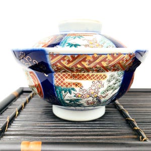 Op de afbeelding: Een Japanse keramische kom met deksel, versierd met ingewikkelde handgeschilderde ontwerpen. De kom toont een levendige mix van kleuren, waaronder blauw, rood, goud en groen, met bloemen- en geometrische patronen. Het staat op een zwarte bamboe lade.
