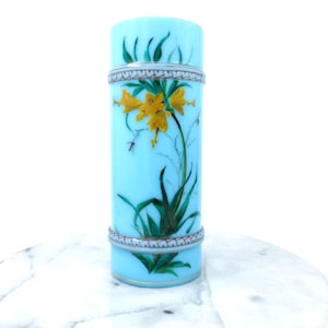 Op de afbeelding: Cilindervormige, lichtblauwe glazen vaas met een handgeschilderd bloemmotief. De vaas heeft gele bloemen, groene bladeren en een decoratieve band rond de boven- en onderkant. De vaas is ongeveer 20 cm hoog.