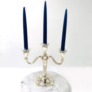 Op de afbeelding: Een zilveren kandelaar met drie marineblauwe kaarsen. De kandelaar heeft een decoratieve voet en gebogen armen die de kaarsen vasthouden. De kaarsen zijn ongeveer 25 cm hoog.