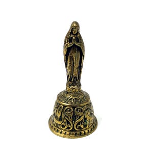 May include: A vintage brass bell with a statue of a robed figure in prayer. The bell has ornate floral and scrollwork designs. The base of the bell has the word "DENNE" and other text. The bell is made of brass.