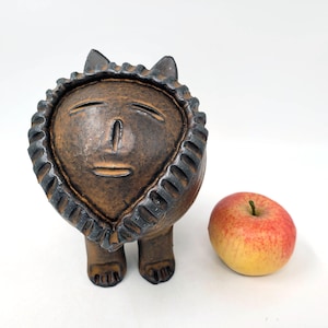 May include: A brown ceramic sculpture of a stylized human face with closed eyes and a small mouth. The sculpture has a textured, heart-shaped head and two small legs. The sculpture is standing on a white surface next to a red apple.