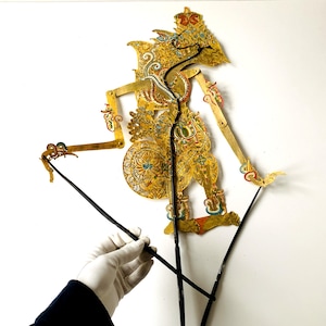 Op de afbeelding: Een gouden Wayang Kulit-pop met ingewikkelde uitgesneden patronen en een kroon. De pop wordt vastgehouden door zwarte staven. Het ontwerp bevat rode, blauwe en witte accenten. De achtergrond is wit.