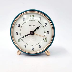 Op de afbeelding: Een teal en gouden analoge wekker met een witte wijzerplaat. De wijzerplaat heeft zwarte wijzers, gouden accenten en het woord "ROSTOV". De klok heeft goudkleurige poten en een klein handvat aan de bovenkant.